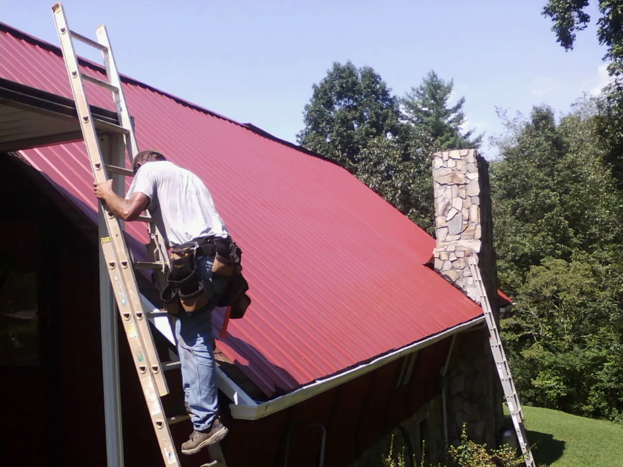 Roofer on a ladder beside a red metal roof and stone chimney — Soffit & Fascia Repair by Roofing Brothers in Pipersville
